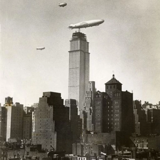Historic photo of the Empire State Building under construction in 1930, surrounded by traditional 19th-century buildings and airships in the sky, illustrating the juxtaposition of the Art Deco skyscraper's futuristic design with its contemporaries.